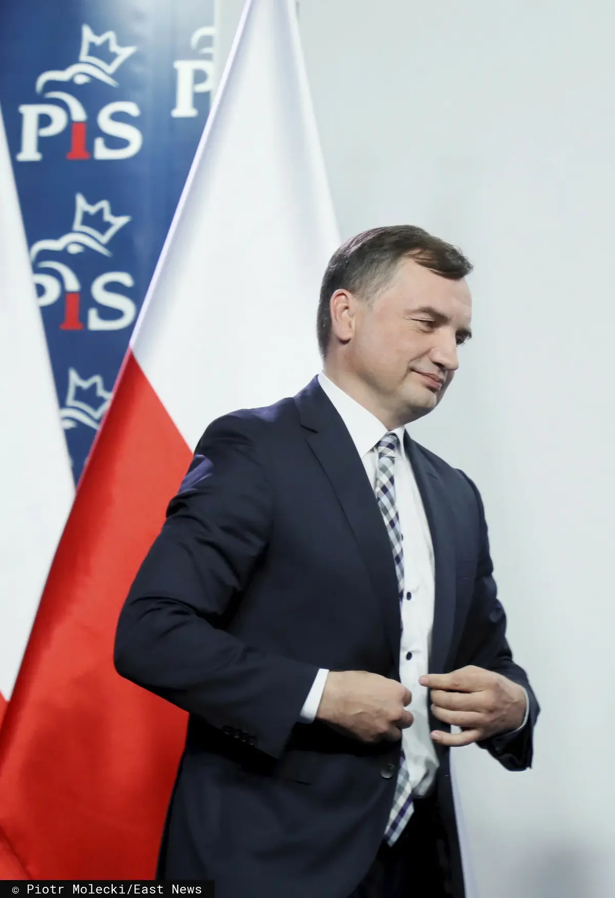 Zbigniew Ziobro zapinający marynarkę na tle flagi Polski i ścianki z logo PiS
