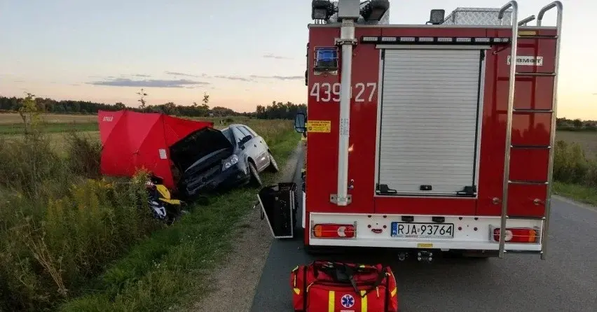 wóz strażacki na szosie, obok samochód po wypadku w rowie i policyjny czerwony parawan