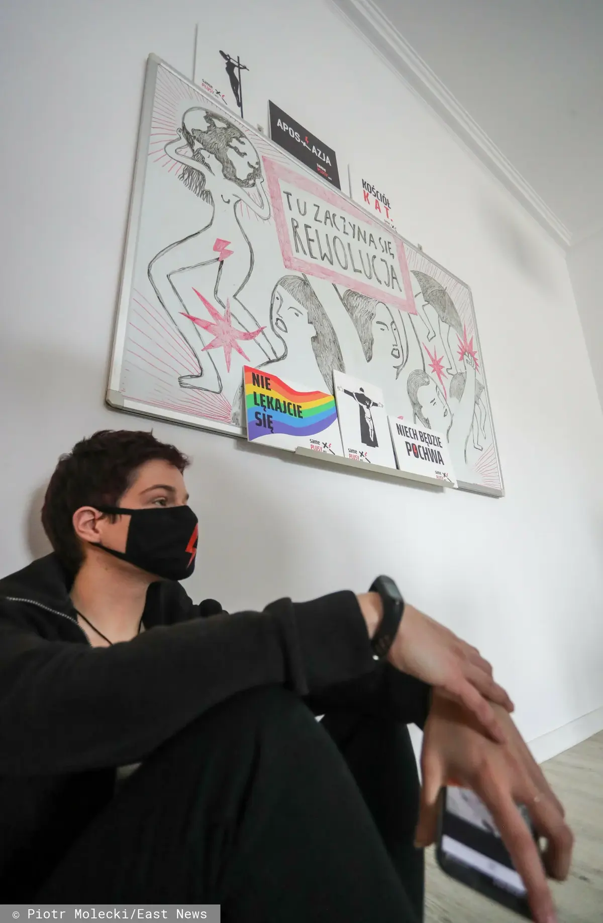 Chłopak ubrany na czarno siedzi na podłodze, w tle plakat z napisem "tu zaczyna się rewolucja"
