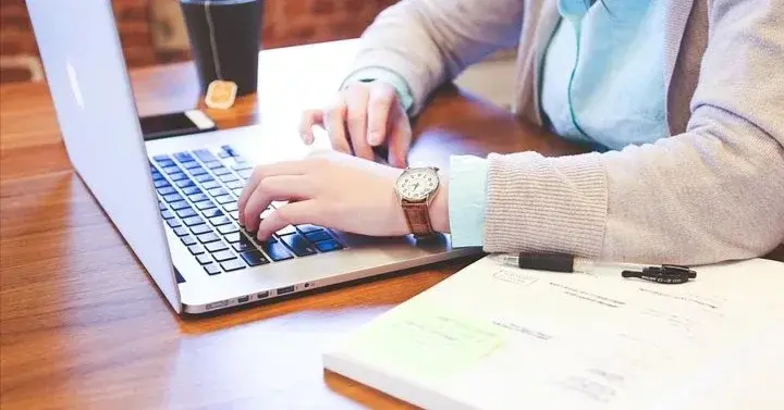 Kobieta pracuje przy laptopie, po jej lewej stronie leży książka, a po prawej kubek z herbatą