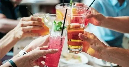 Cztery osoby trzymają w dłoniach szklanki z napojami, siedząc nad zastawionym jedzeniem stołem