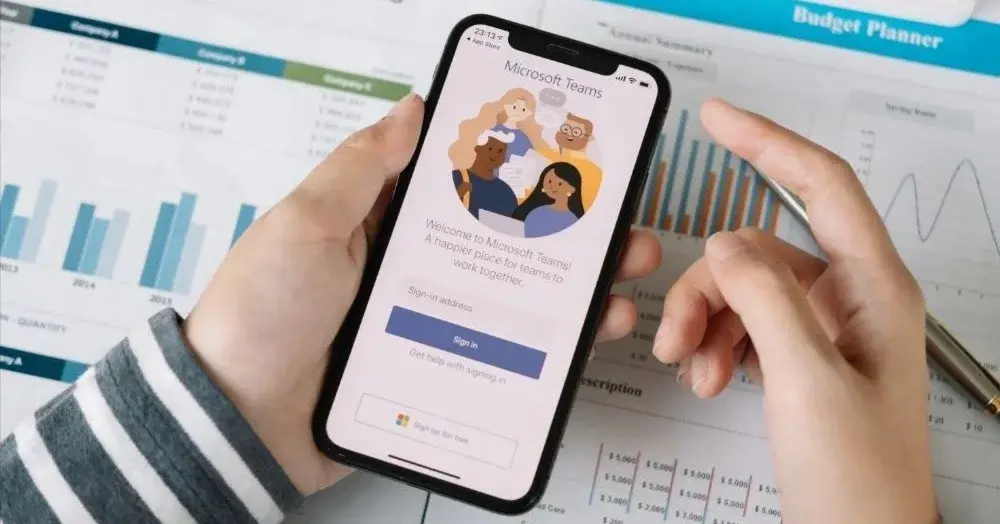 Dłonie trzymające smartfon z otwartą aplikacją Microsoft Teams na tle dokumentów