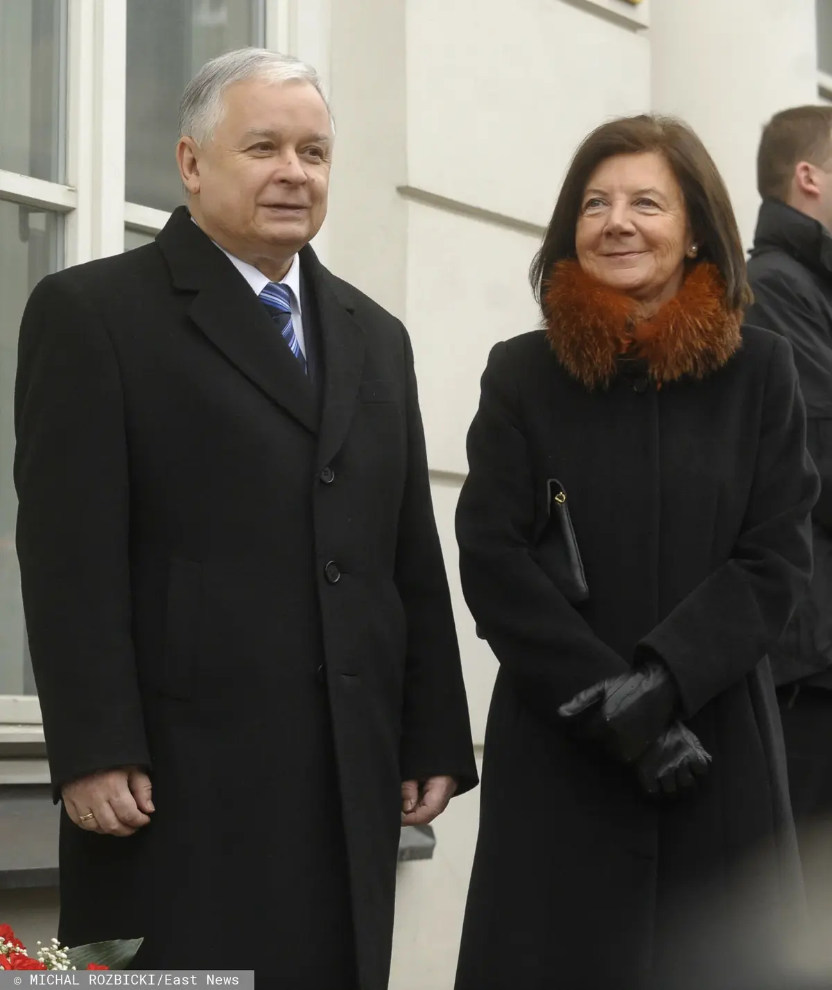 Lech Kaczyński w czarnym płaszczu i białej koszuli z krawatem obok Marii Kaczyńskiej ubranej w czarny płaszcz z pomarańczowo-czerwonym futrem przy szyi