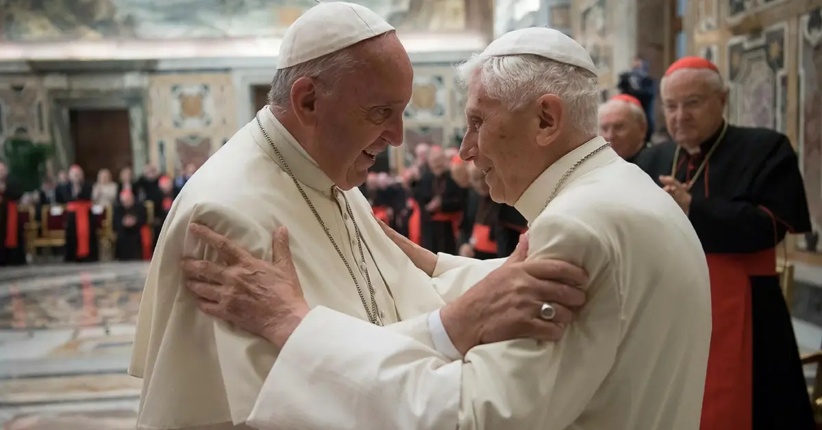 papież franciszek obejmuje się z papieżem benedyktem XVI w tle widać grupę arcybiskupów 