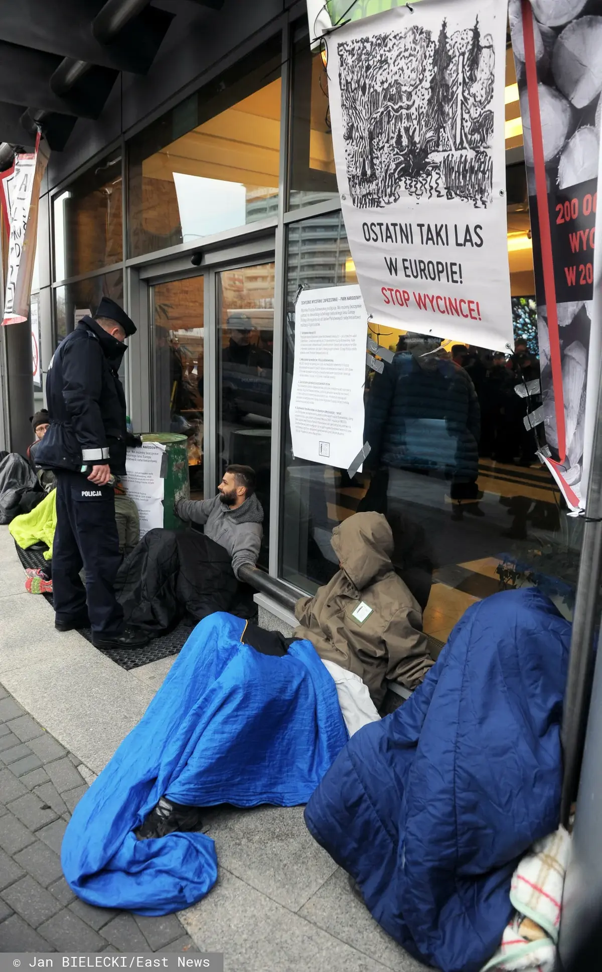 Osoby protestujące i policjant pod siedzibą Lasów Państwowych w Warszawie na tle plakatu z napisem "Ostatni taki las w Europie! Stop wycince!"