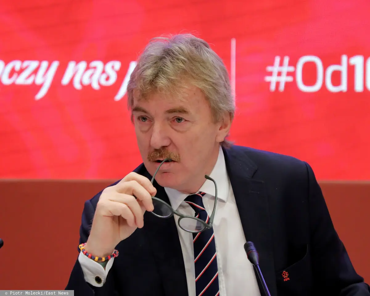 Zbigniew Boniek na czerwonym tle podczas konferencji prasowej przygryzający okulary