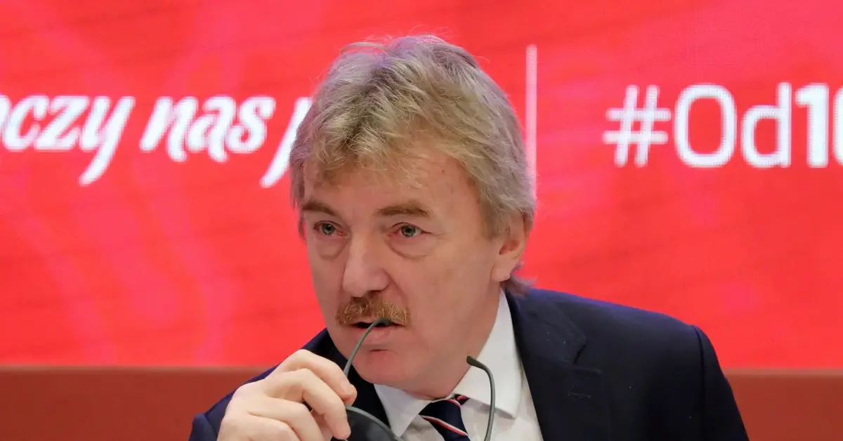 Zbigniew Boniek na czerwonym tle podczas konferencji prasowej przygryzający okulary