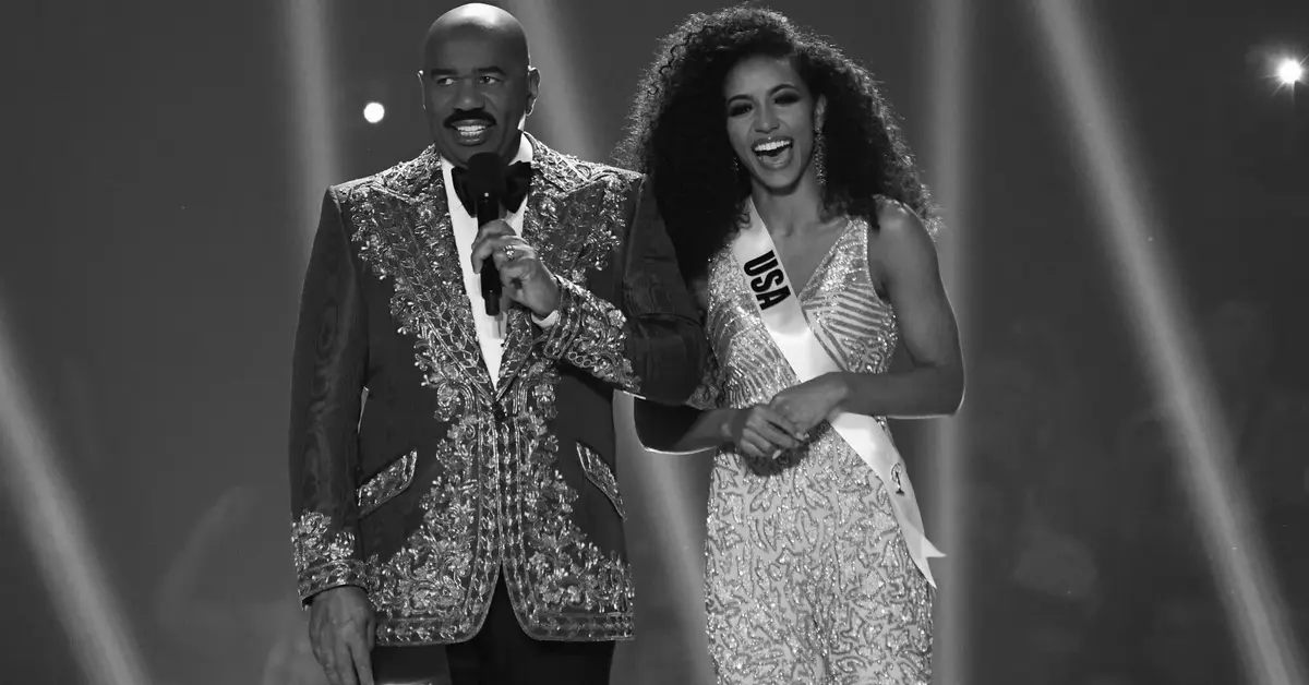 Cheslie Kryst na scenie w wieczornej kreacji z szarfą miss USA obok uśmiechniętego Stevea Harveya