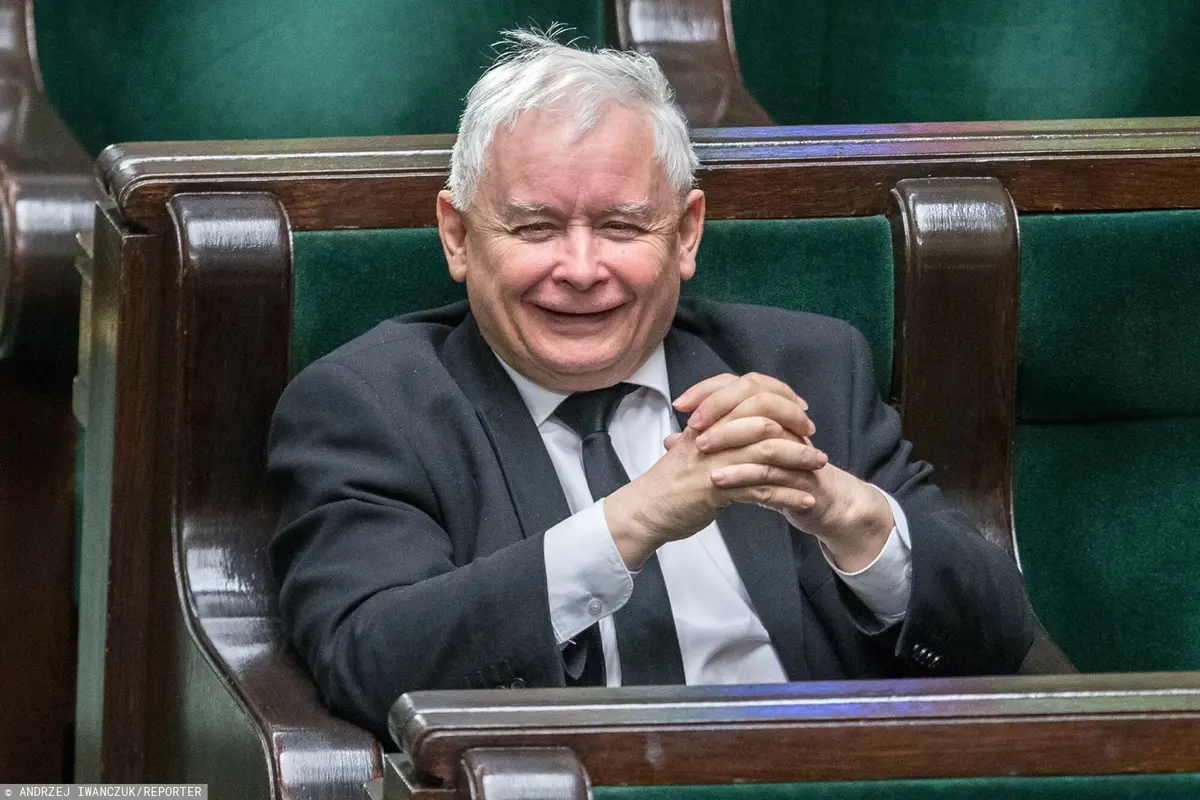 Uśmiechający się Jarosław Kaczyński ze złożonymi rękoma rozparty w swoim miejscu poselskim na sali plenarnej Sejmu 