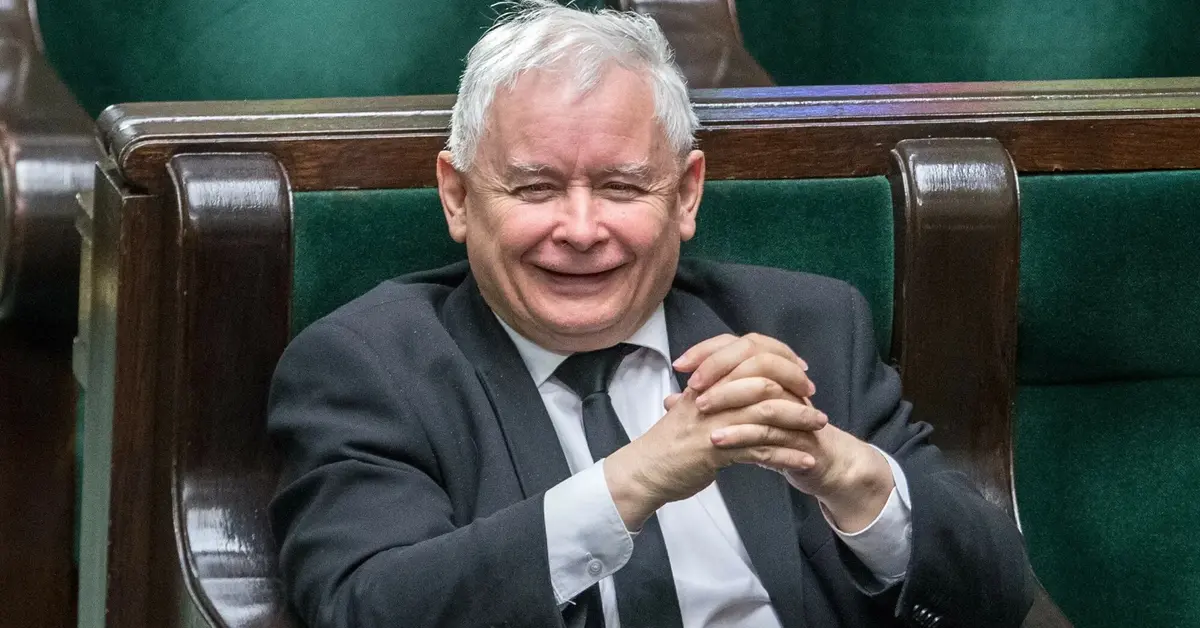 Uśmiechający się Jarosław Kaczyński ze złożonymi rękoma rozparty w swoim miejscu poselskim na sali plenarnej Sejmu 