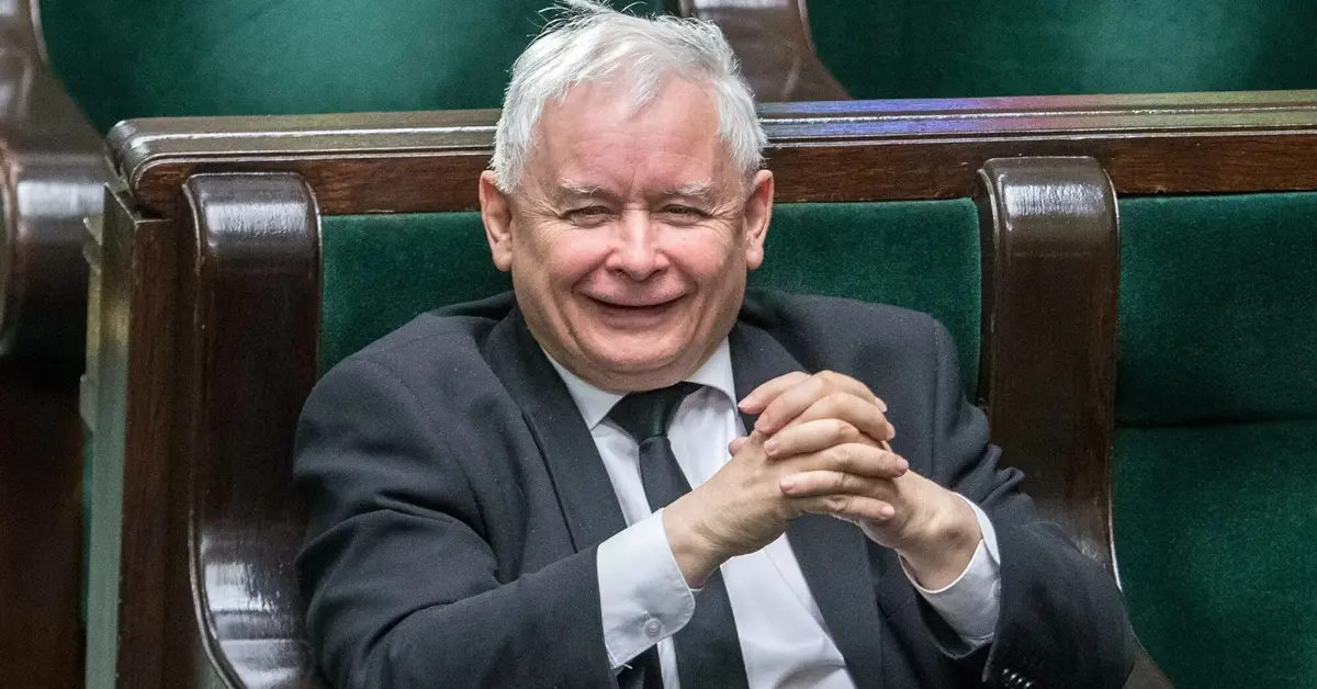 Uśmiechający się Jarosław Kaczyński ze złożonymi rękoma rozparty w swoim miejscu poselskim na sali plenarnej Sejmu 