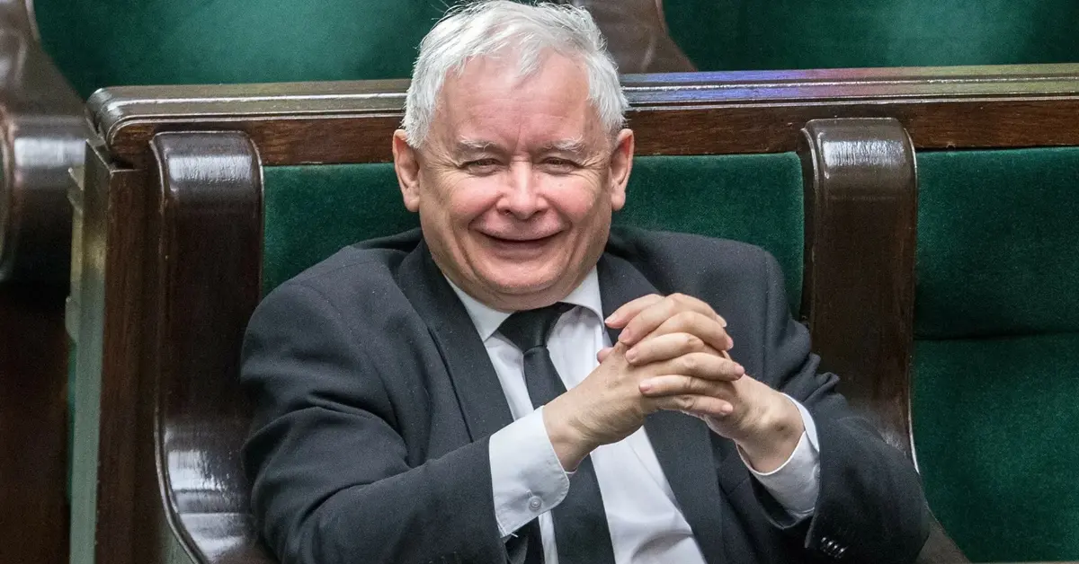 Uśmiechający się Jarosław Kaczyński ze złożonymi rękoma rozparty w swoim miejscu poselskim na sali plenarnej Sejmu 