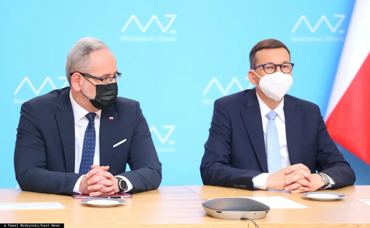 adam niedzielski siedzi za stołem podczas konferencji i patrzy w stronę siedzącego obok mateusza morawieckiego