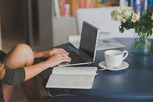 Kobieta siedzi przy laptopie, a obok na stole leży zeszyt i filiżanka