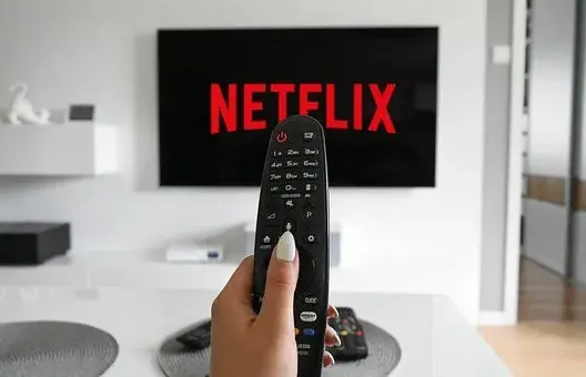 Osoba trzyma w dłoni pilota, w tle znajduje się ekran z napisem Netflix