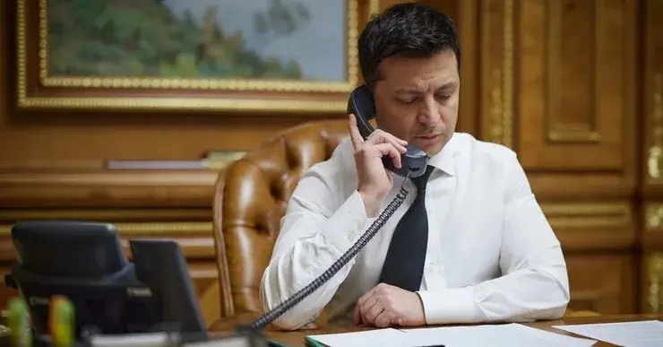 Wołodymyr Zełenski siedzi w gabinecie przy biurku na skórzanym fotelu i trzyma przy uchu słuchawkę telefonu stacjonarnego