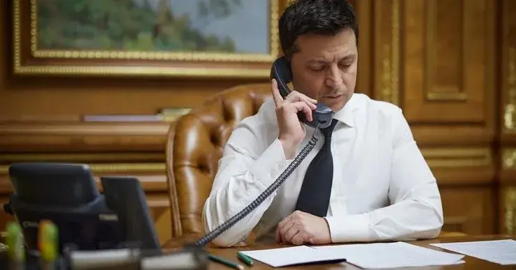 Wołodymyr Zełenski siedzi w gabinecie przy biurku na skórzanym fotelu i trzyma przy uchu słuchawkę telefonu stacjonarnego