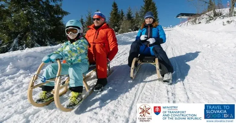 Tata z dzieckiem obok mama, wszyscy w zimowych kolorowych skafandrach,  na sankach na górce w słowackich Pieninach