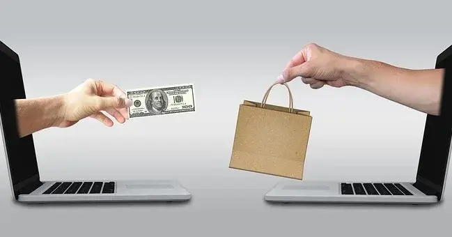 Dwa laptopy, z każdego z nich wychodzi ludzka ręka - jedna podaje pieniądze, a druga torbę z zakupami