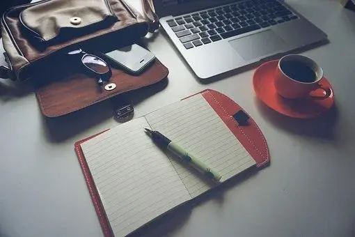 Biurko, na którym leży aktówka, laptop, filiżanka z czarną kawą, zeszyt i długopis