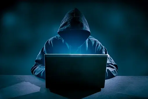 Osoba w kapturze siedzi przed laptopem, wyglądając jak haker w trakcie cyberataku