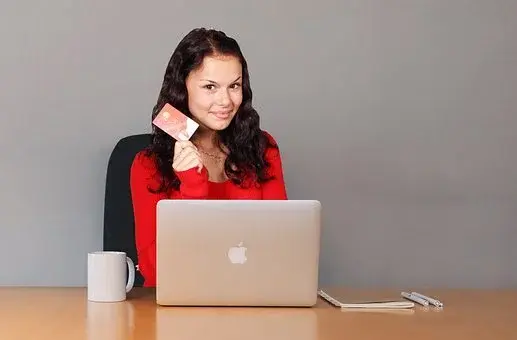 Kobieta w ciemnych włosach i czerwonej bluzce, siedzi przed laptopem marki Apple, a w dłoni trzyma kartę kredytową