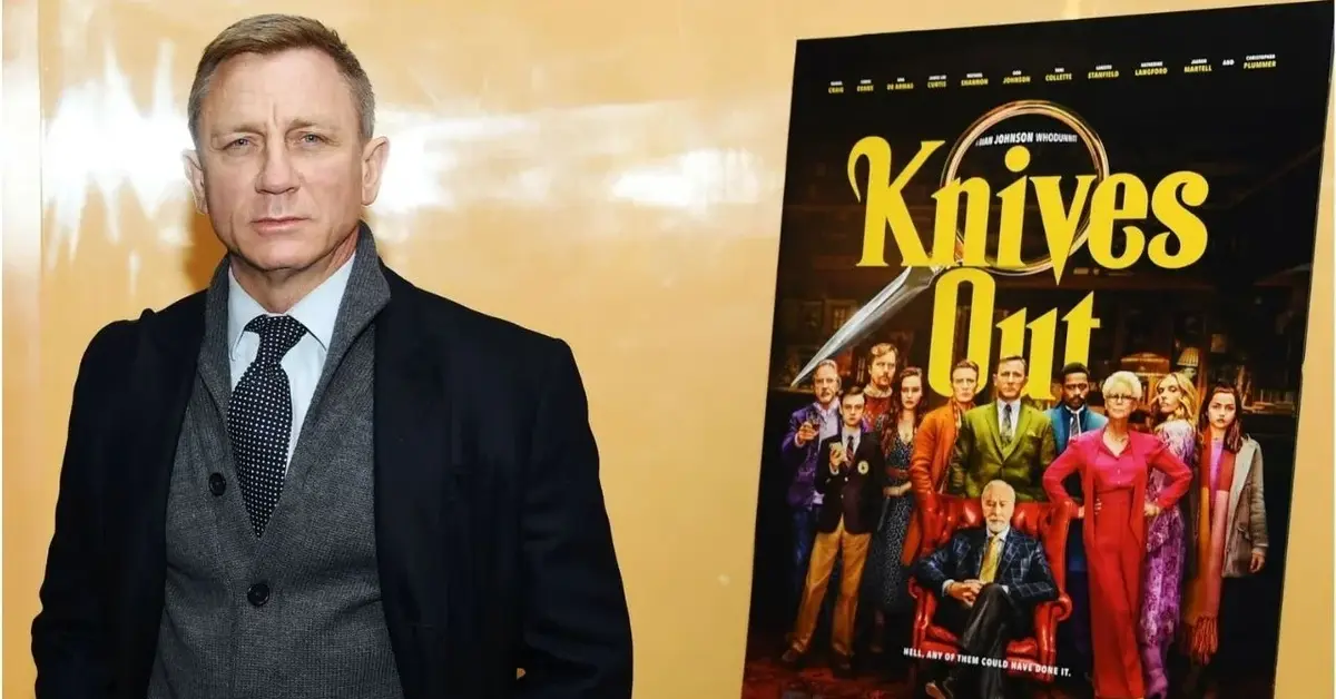 Daniel Craig w szarym swetrze i granatowej marynarce obok plakatu filmu Riana Johnsona "Na noże"
