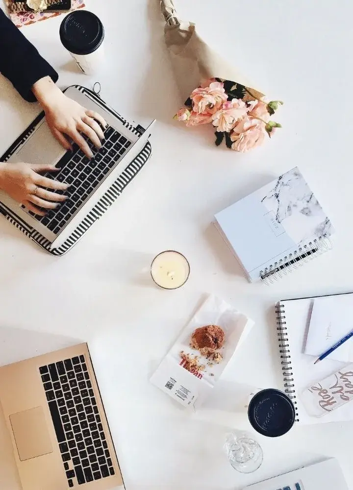 Rzut z góry na białe biurko z dwoma laptopami, kwiatami i notatkami