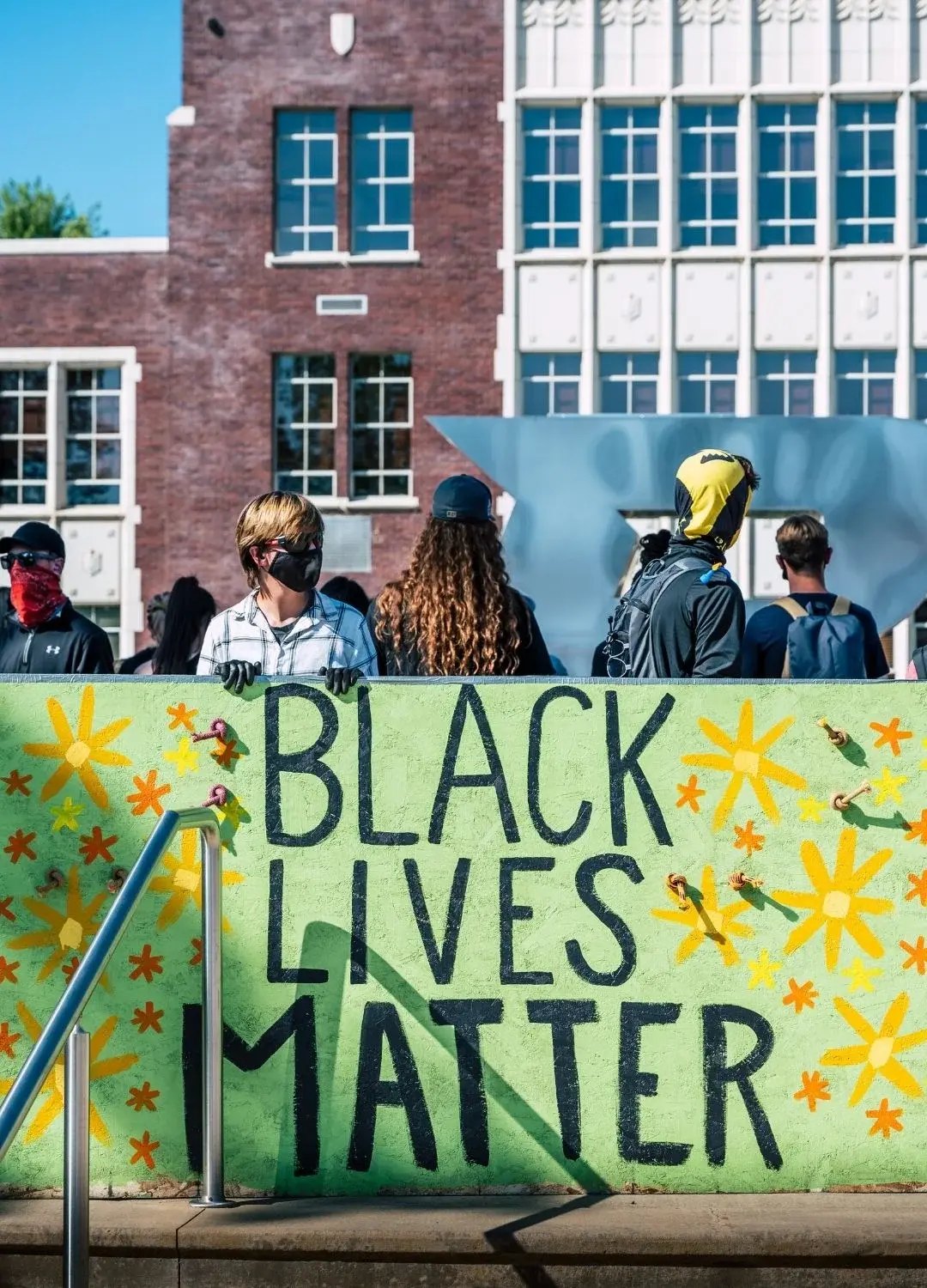 Zielony transparent z napisem "Black Lives Matter" przed osobami protestującymi na tle budynku