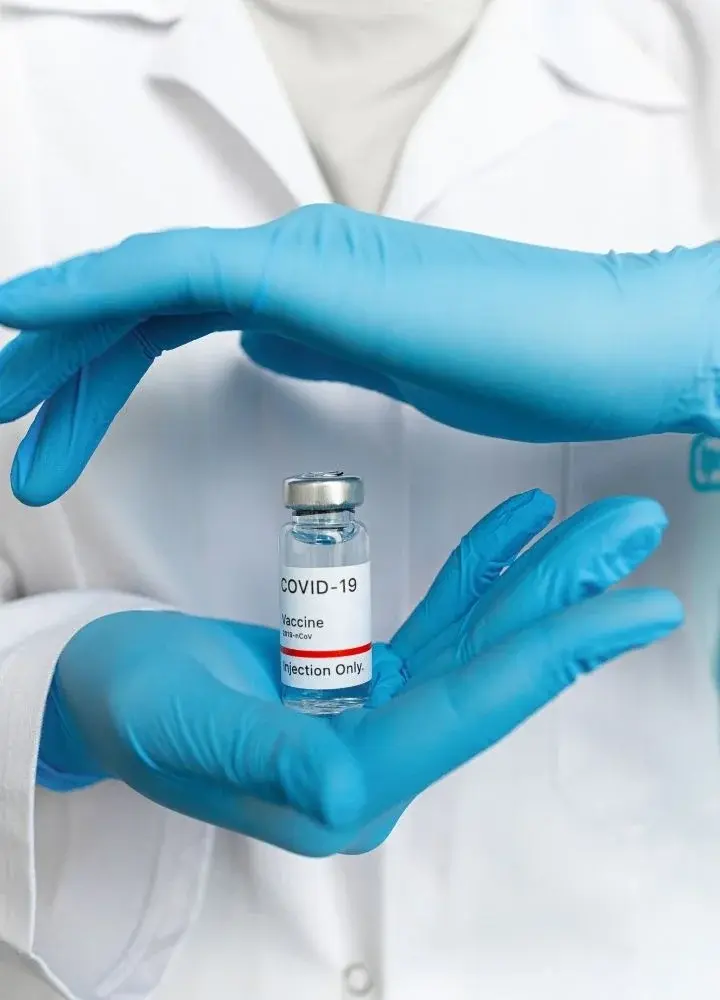 Dłonie w błękitnych rękawiczkach trzymające fiolkę ze szczepionką na COVID-19 na tle białego fartucha lekarskiego