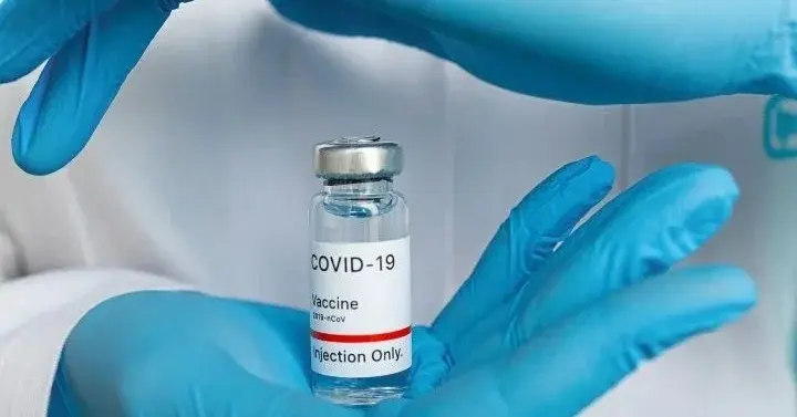 Dłonie w błękitnych rękawiczkach trzymające fiolkę ze szczepionką na COVID-19 na tle białego fartucha lekarskiego