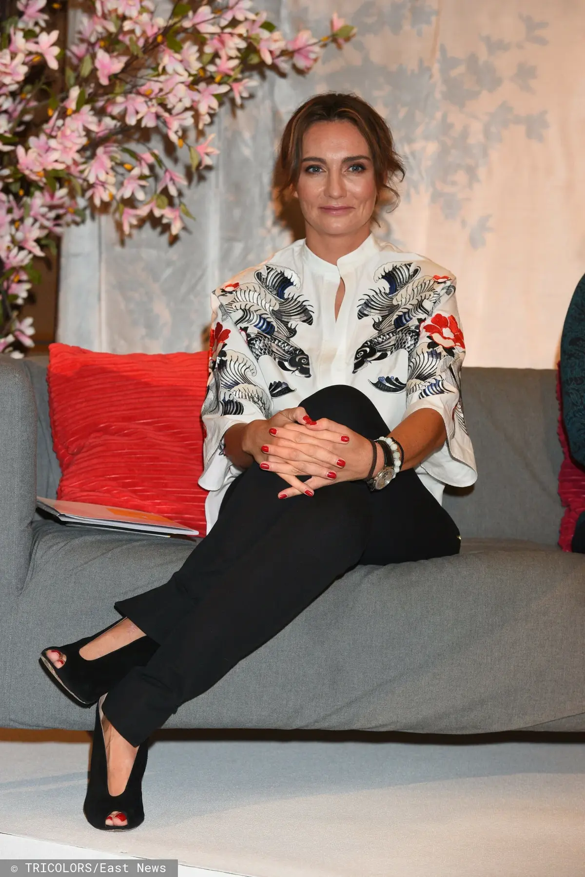 Dominika Kulczyk w ciemnych spodniach i bluzce w kwiaty na kanapie