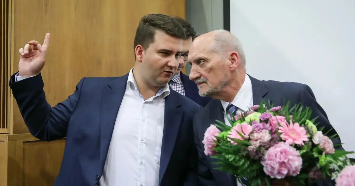 Bartłomiej Misiewicz pokazuje coś Antoniemu Macierewiczowi, który trzyma bukiet kwiatów 