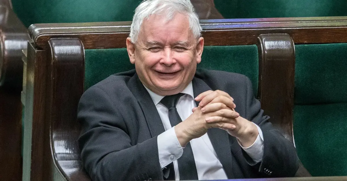 Uśmiechający się Jarosław Kaczyński ze złożonymi rękoma rozparty w swoim miejscu poselskim na sali plenarnej Sejmu 