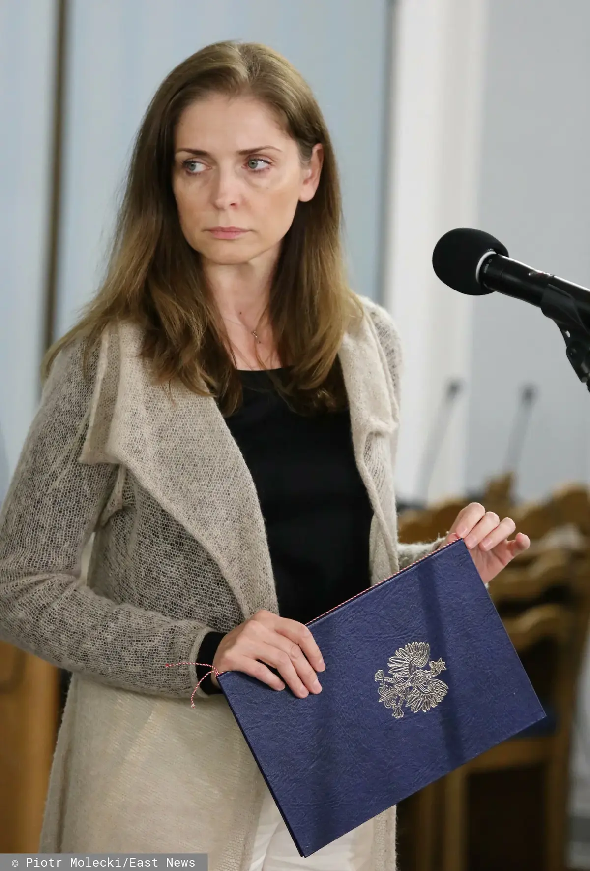 Joanna Mucha w szaro-beżowym płaszczu stojąca przy mikrofonie, trzymając w ręku granatową teczkę z godłem Polski