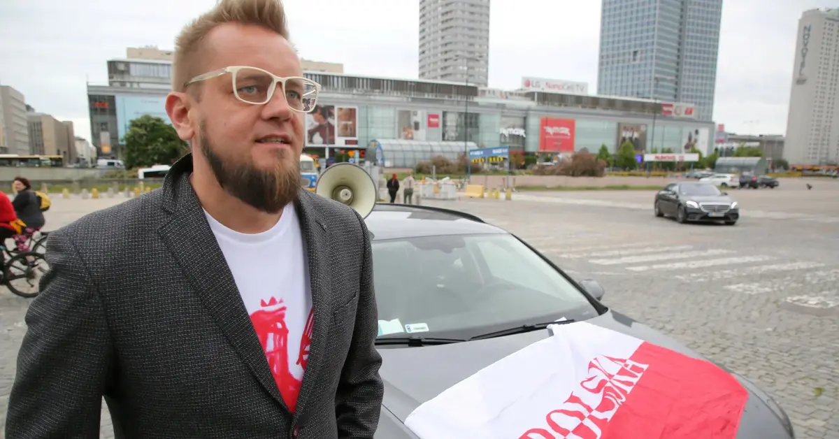 Paweł Tanajno obok samochodu z polską flagą.