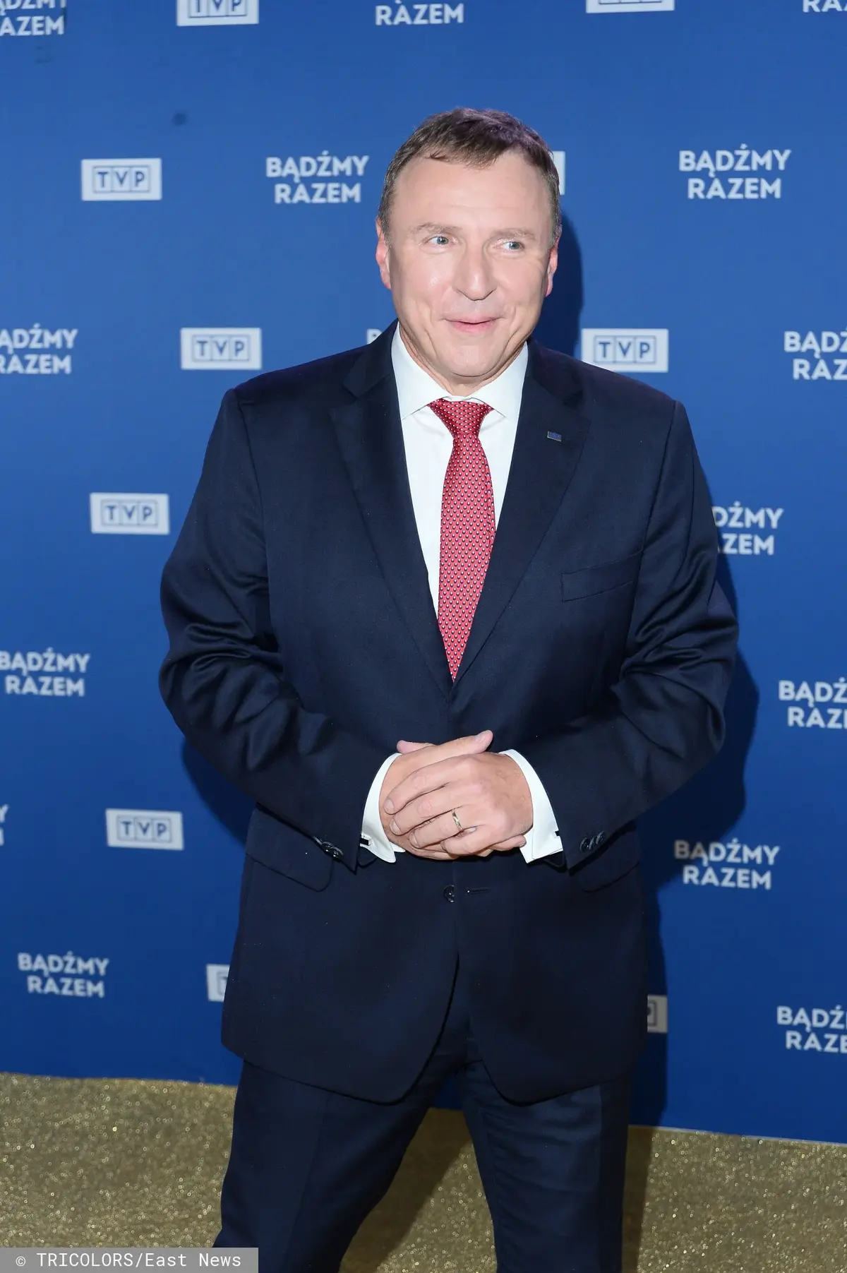 Uśmiechnięty Jacek Kurski w granatowym garniturze, białej koszuli i czerwonym krawacie na tle niebieskiej ścianki z logo TVP
