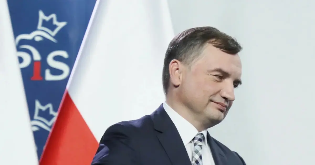 Zbigniew Ziobro zapinający marynarkę na tle flagi Polski i ścianki z logo PiS
