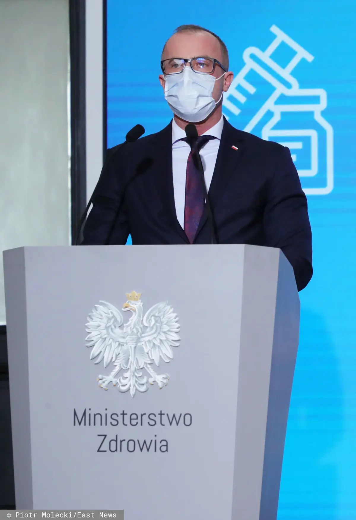Grzegorz Cessak w garniturze i maseczce za mównicą z napisem Ministerstwo Zdrowia