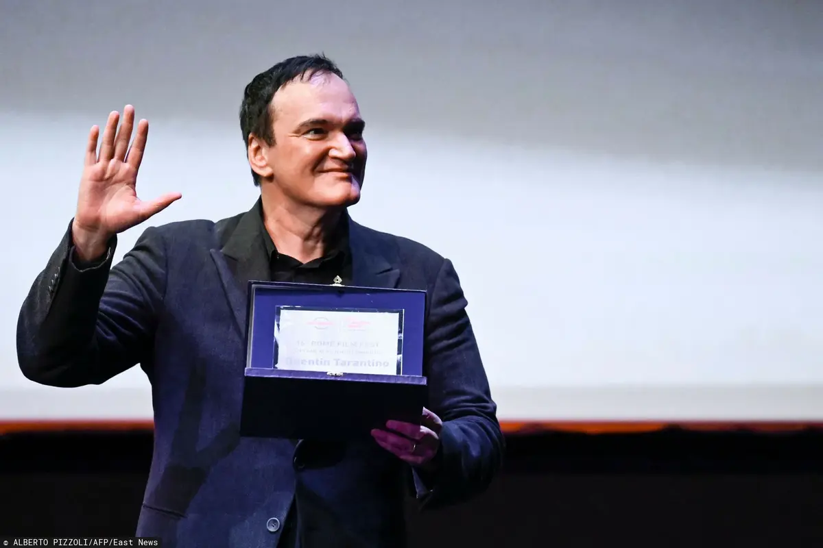 Quentin Tarantino w garniturze pozuje z nagrodą i macha do widzów.