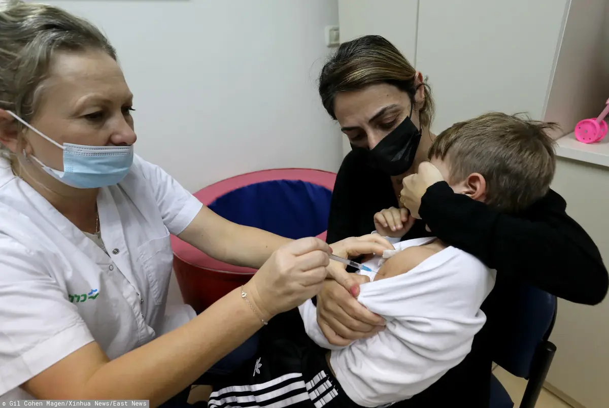 Pielęgniarka w maseczce robi zastrzyk przerażonemu chłopcu, którego trzyma matka, zasłaniając mu oczy