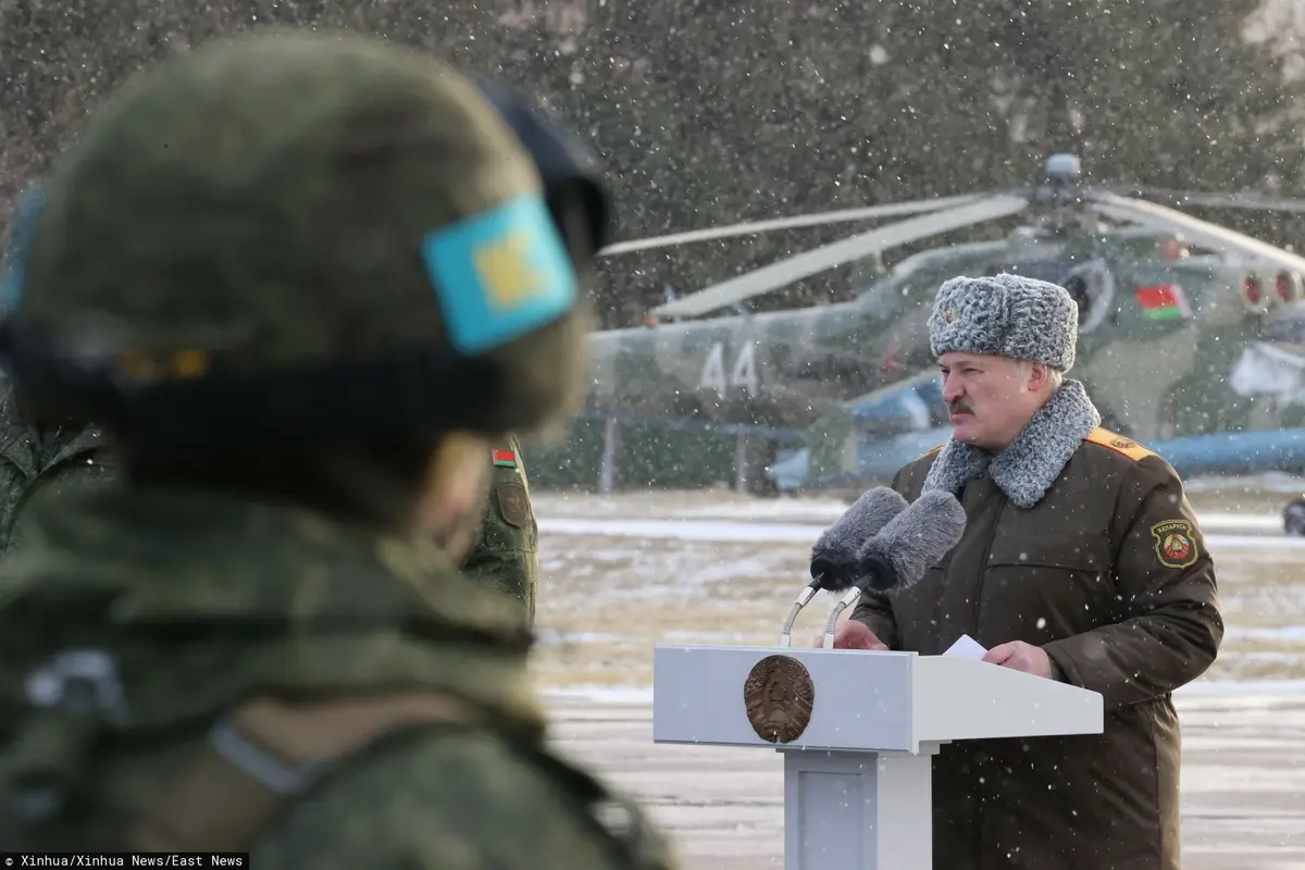 łukaszenka przemawia w mundurze i zimowej czapie za pulpitem, w tle jest helikopter wojskowy, a z przodu żołnierz
