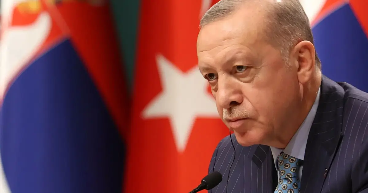 erdogan przed mikrofonem z groźną miną na tle tureckiej flagi