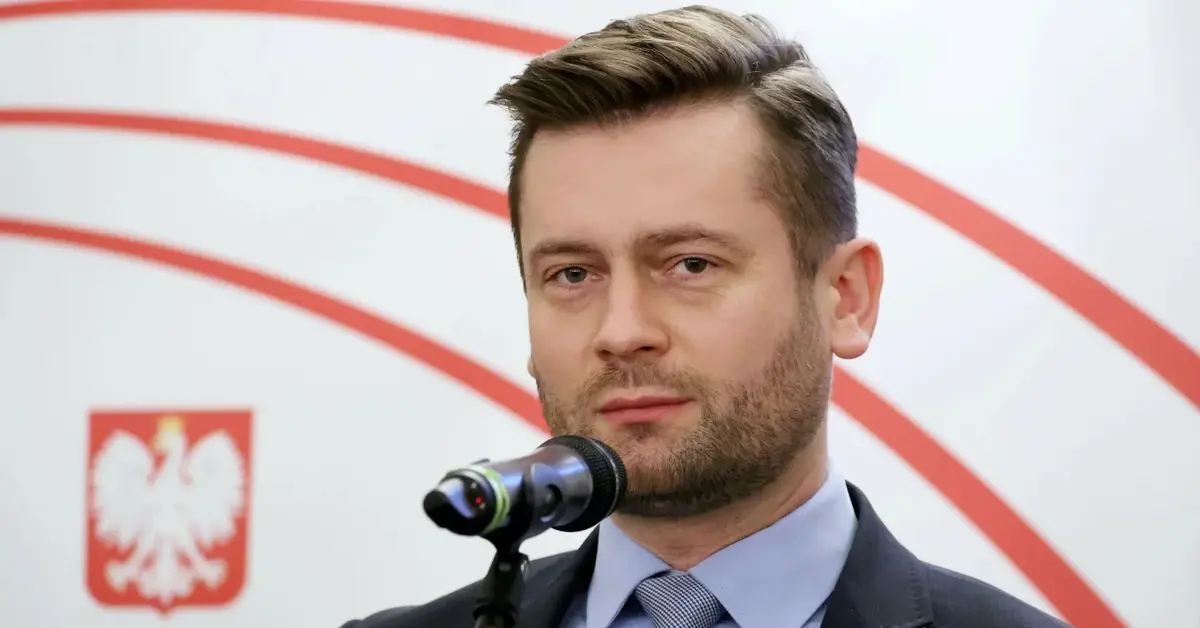 Kamil Bortniczuk stoi w garniturze przed mikrofonem, a w tle m.in. polskie godło