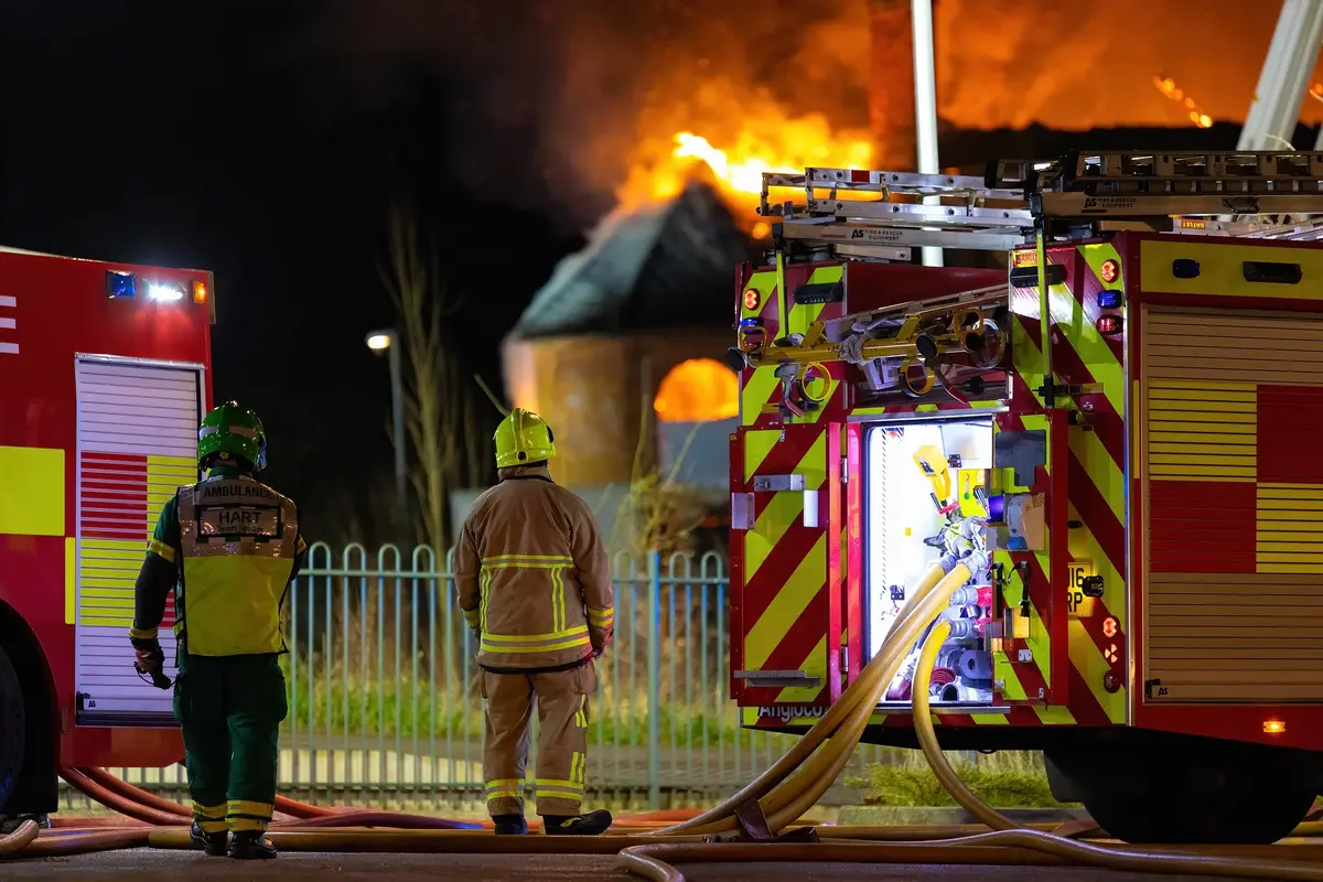 dwaj strażacy stoją przy wozach gaśniczych, a w tle widać płonący budynek 