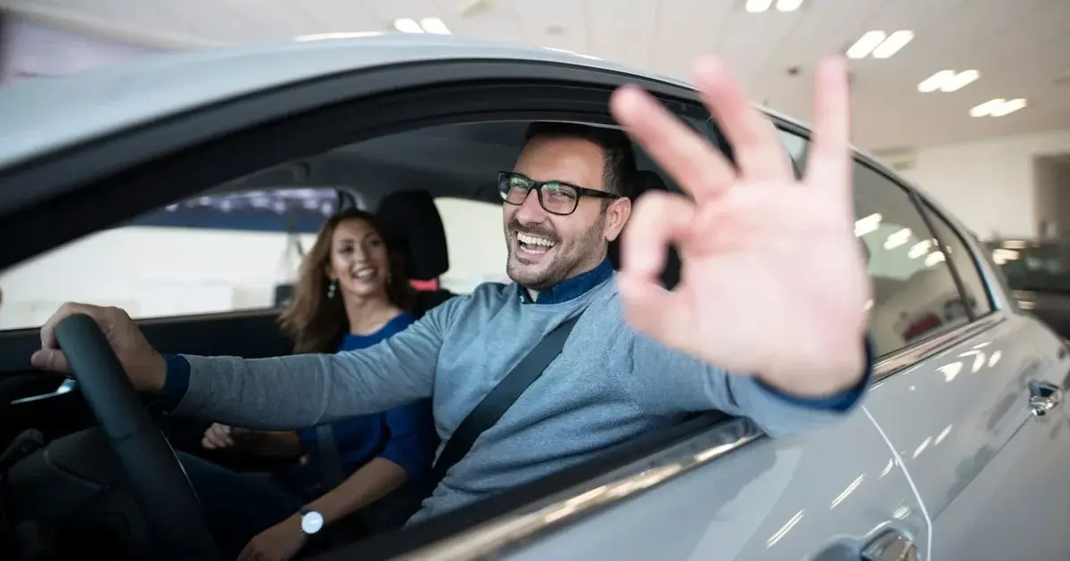 W sali wystawowej srebrny samochód osobowy, z uśmiechniętą pasażerką oraz kierowcą w okularach pokazującym znak OK