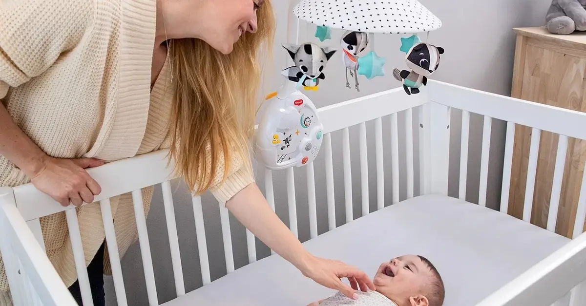 Uśmiechnięta matka pochylona nad dzieckiem w białym łóżeczku z karuzelą do łóżeczka