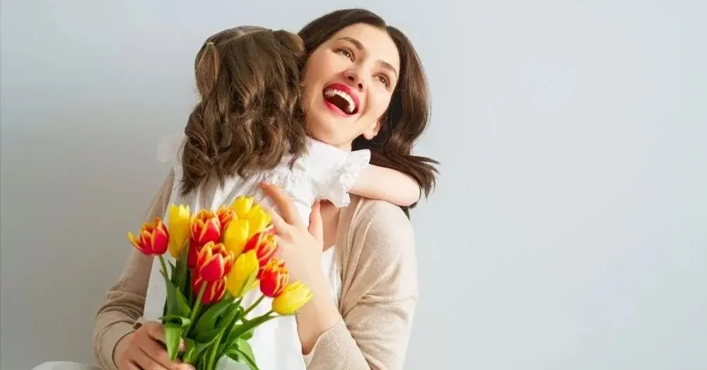Szeroko uśmiechnięta matka z kwiatami w ręku, którą przytula córka