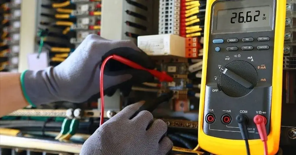 Uruchomiony miernik do prądu na tle instalacji elektrycznych i dłoni w rękawiczkach ochronnych