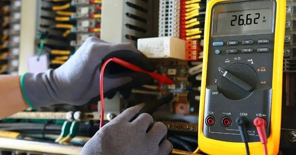Uruchomiony miernik do prądu na tle instalacji elektrycznych i dłoni w rękawiczkach ochronnych